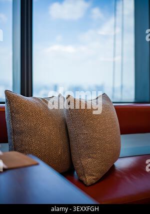 Two patterned grey throw pillows on a red leather bench by a high-rise window overlooking a blurred city skyline. Stock Photo