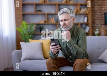 Thoughtful senior man with gray hair and beard sitting on a couch, holding a smartphone and looking confused or perplexed while interacting with techn Stock Photo