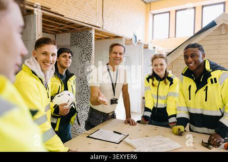 Smiling instructor gesturing while standing with vocational students around workbench inside construction workshop at up Stock Photo