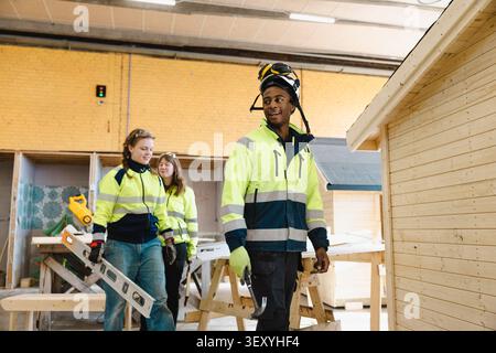 Young male vocational student wearing headphones and hardhat on head walking in workshop towards training session at sch Stock Photo