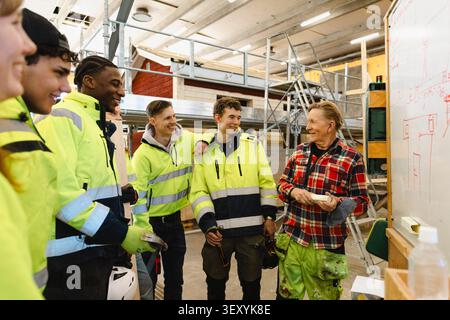 Smiling senior male professor talking with students while standing near whiteboard in workshop at vocational school Stock Photo