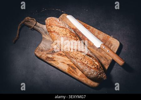 Freshly baked rustic bread baguette with seeds on wooden cutting board with knife Stock Photo