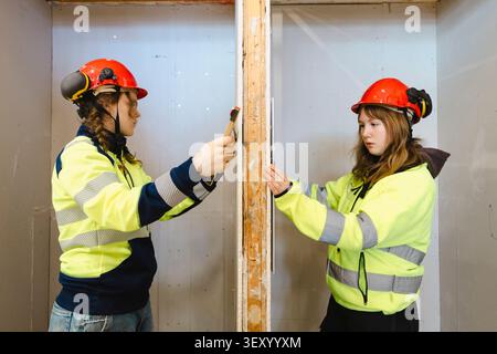 Two female vocational students working together while measuring drywall panel with ruler on opposite sides inside traini Stock Photo