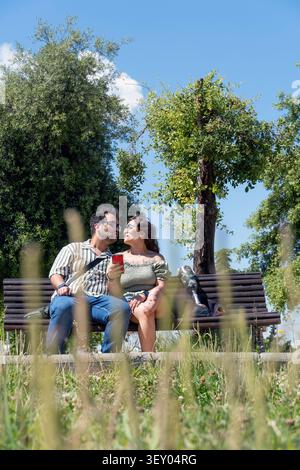 Loving couple sitting on a park bench, the man having a leg prosthesis, enjoying a sunny day outdoors, sharing a moment Stock Photo