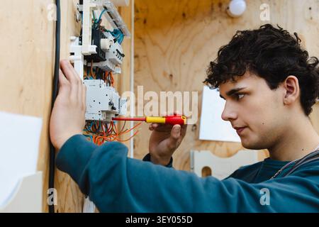 Focused vocational male student using screwdriver on complex training circuit board breaker assembly in workshop at scho Stock Photo