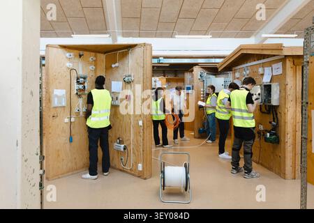 Young vocational students engaged in hands on tasks inside separate electrical training booths within large instructiona Stock Photo