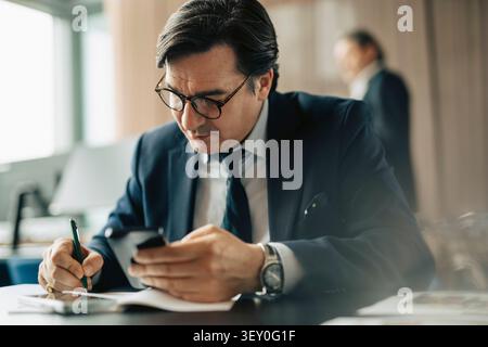 Focused mature male business professional using smart phone while writing on document in office Stock Photo