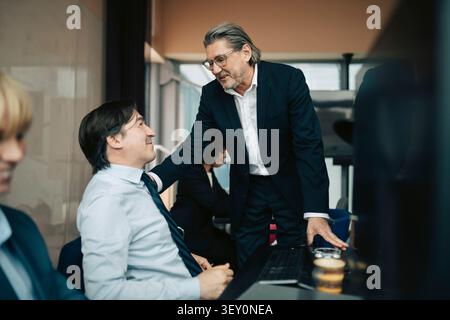 Senior businessman talking with hand on shoulder of male colleague sitting at desk in office Stock Photo