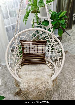 Modern cozy beautiful room with a braided rope macrame chair, green plants and a window with curtains. Interior and background. Location for photo sho Stock Photo