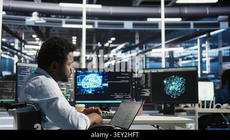 Data infrastructure center computer scientist in wheelchair working with AI visual programming system to minimize system downtime, while with disability working with AI visual programming tool. Stock Photo