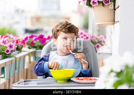 Happy boy having breakfast on a flower‑filled balcony, enjoying a bright and colorful outdoor moment Stock Photo