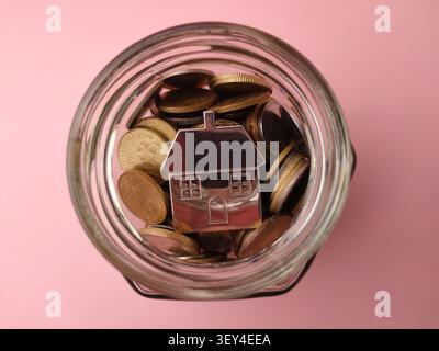 Top down view looking into a glass jar filled with coins and a small metallic house figurine against pink. Stock Photo