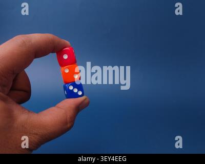 A hand vertically stacks and holds three colorful plastic dice—red, orange, and blue against a blue background. Stock Photo