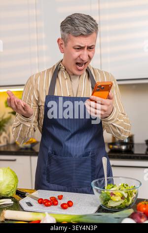 Middle-aged man in kitchen stares at smartphone with pained face after bad results and troubling message. Young adult guy reacts to defeat failure and online problem during home routine with stress. Stock Photo
