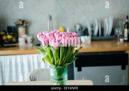 A beautiful arrangement of pink tulips in a stylish green vase, placed on a wooden dining table in a cozy home kitchen interior. Stock Photo