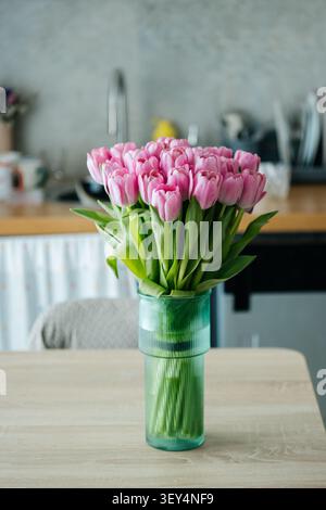 A beautiful arrangement of pink tulips in a stylish green vase, placed on a wooden dining table in a cozy home kitchen interior. Stock Photo