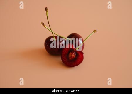 Three dark red cherries with stems on a soft peach background. Simple fruit still life with copy space, fresh summer feel, and clean minimal style. Stock Photo