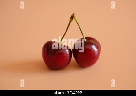 Two ripe red cherries with stems leaning together on a soft peach background. Simple studio fruit still life with copy space, fresh summer mood and cl Stock Photo