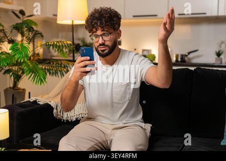 Arabian man at home checks smartphone gets bad news game loss lottery frowns shakes head no sighs. Egyptian guy on sofa reads message about result looks upset frustrated sad reacts to negative alert. Stock Photo