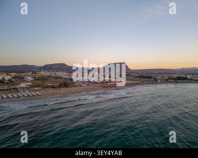 Aerial view displays ocean waves rolling onto sandy beach next to resort buildings and distant mountains at sunset. Stock Photo