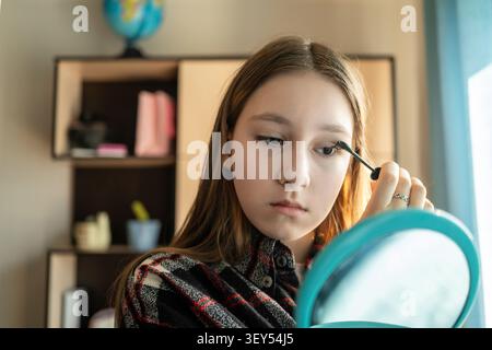 Girl in a plaid shirt applying mascara to her eyelashes in front of a round blue mirror. Focus on makeup, daily beauty routine, natural daylight. Stock Photo