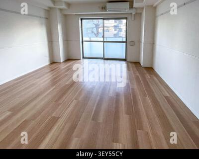 An empty apartment living room in Japan features light wood laminate flooring, white walls, and a sliding glass balcony door letting in soft natural light. Stock Photo