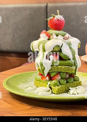 A tall stack of matcha green tea pancakes topped with cream sauce, strawberries, and green cream sits on a green plate on a wooden table, with a gray sofa booth visible in the background. Stock Photo