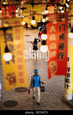 Crowd Walking on Pedestrian Street Behind Blurred Yellow Banners. Delivery Driver Walking in Blue Uniform on Busy Sanya Street Stock Photo