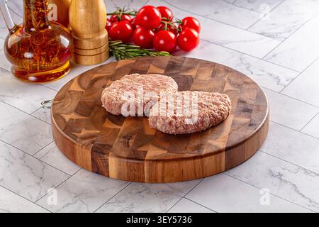 Two raw meat patties on round wooden cutting board. Background features cherry tomatoes, rosemary, olive oil bottle, pepper mill. Marble countertop, w Stock Photo