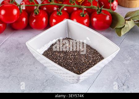 Chia seeds fill textured white bowl against gray stone surface. Vibrant red cherry tomatoes on vine dried bay leaves add color contrast. Soft natural Stock Photo