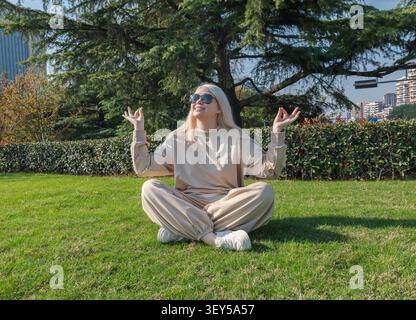 Blonde woman sitting cross-legged in a city park meditating under a sunny sky on a bright afternoon Stock Photo