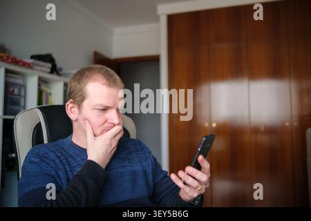 Man holding smartphone with hand on face, thinking at his home office desk Stock Photo