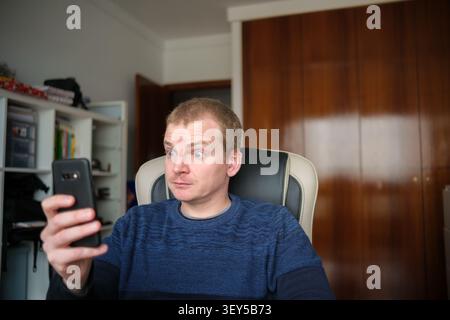 Man at home reacting with wide eyes and open mouth to shocking news on a smartphone Stock Photo