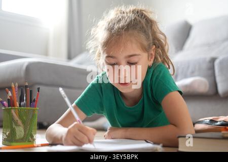 Elementary School Girl Writing Homework While Lying on Living Room Floor at Home Stock Photo