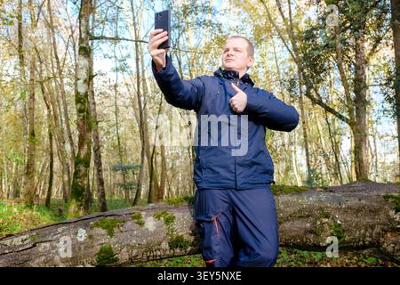 Man taking a selfie with a smartphone, leaning on a log in a forest, giving a thumbs up Stock Photo