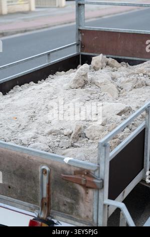 Weathered open metal trailer packed with coarse sand, rubble and plaster debris parked beside urban curb under soft daylight, showing rusted sidewalls Stock Photo