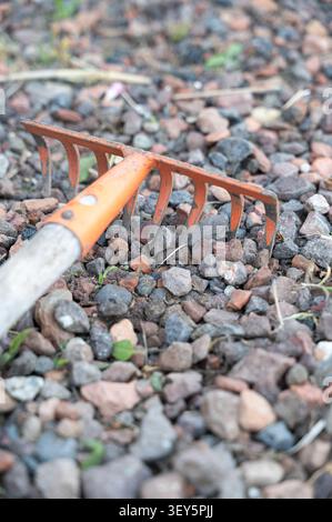 Rusty garden rake resting on multicolored gravel with scattered leaves, weathered metal tines, wooden handle, and rough stone texture creating Stock Photo