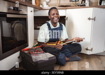 Young female plumber laughing while fixing pipes under kitchen sink Stock Photo