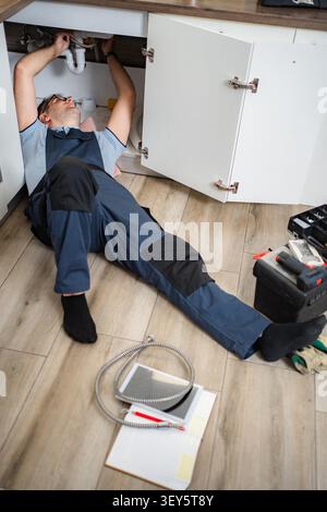 Plumber lying on floor repairing kitchen sink drainage system Stock Photo