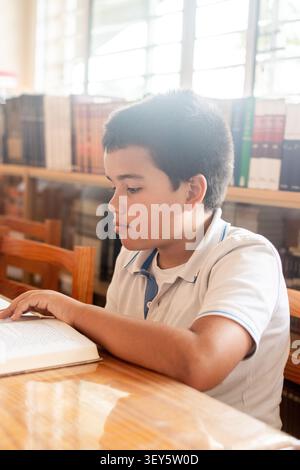 Focused elementary school boy reading a book while sitting at a table in a bright school library. Stock Photo