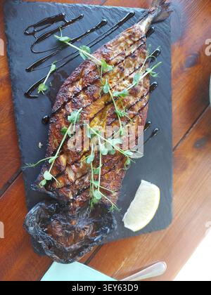Grilled fish served on a slate plate, in wooden table, with grilled fish, lemon wedge, herbs, dark sauce, with slate plate, wooden table texture, from a top-down view perspective, clear detail. Stock Photo