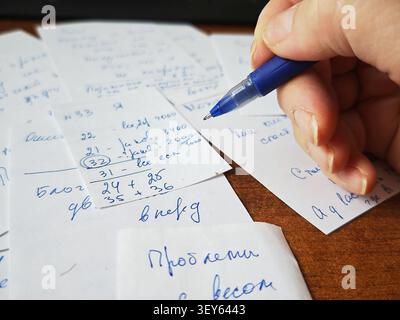 Kirov, Russia - February 24, 2026: A hand writes on a piece of paper while many small notes with written text are spread on a wooden table surface. Stock Photo