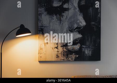 Warm light casts gentle glow on wall. Abstract face art features bold, textured strokes. Expression conveys calm introspection, quiet mood. Modern lam Stock Photo