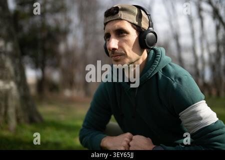 Man wearing headphones and hoodie looking thoughtful while resting outdoors in a park Stock Photo