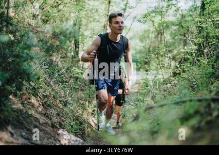 Young man hiking uphill on a forest trail with backpack and trekking poles Stock Photo