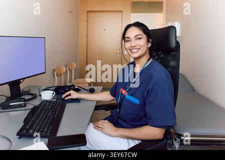 Portrait of female medical professional in blue scrubs smiling while sitting at her office desk Stock Photo