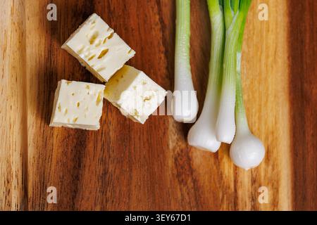 A macro top-view photograph of three diced cubes of white feta cheese and fresh, wet green spring onions lying on a textured wooden cutting board. The Stock Photo