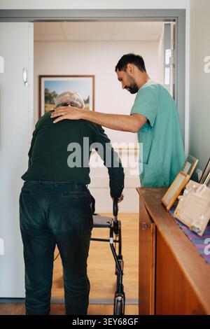 Male healthcare worker supporting old man walking with support of walker at retirement home Stock Photo