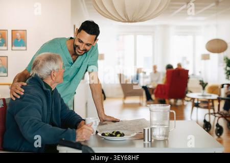 Smiling male nurse interacting with senior man sitting at table in retirement home Stock Photo