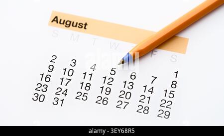 An orange ballpoint pen lying diagonally on a white paper calendar page for the month of August. Close up. Stock Photo